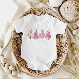 K&ouml;nnte beinhalten: Wei&szlig;er Baby-Body mit einem Design von vier rosa Weihnachtsb&auml;umen. Die B&auml;ume haben verschiedene Rosat&ouml;ne und dekorative Elemente. Der Body wird in einem Weidenkorb pr&auml;sentiert.