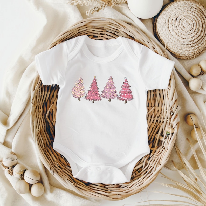 K&ouml;nnte beinhalten: Wei&szlig;er Baby-Body mit einem Design von vier rosa Weihnachtsb&auml;umen. Die B&auml;ume haben verschiedene Rosat&ouml;ne und dekorative Elemente. Der Body wird in einem Weidenkorb pr&auml;sentiert.