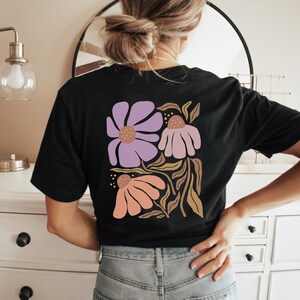 May include: A black short-sleeve t-shirt featuring a large graphic on the back. The design displays three stylized flowers in shades of lavender, light pink, and peach, accompanied by brown leafy stems and small yellow dots on a black background.