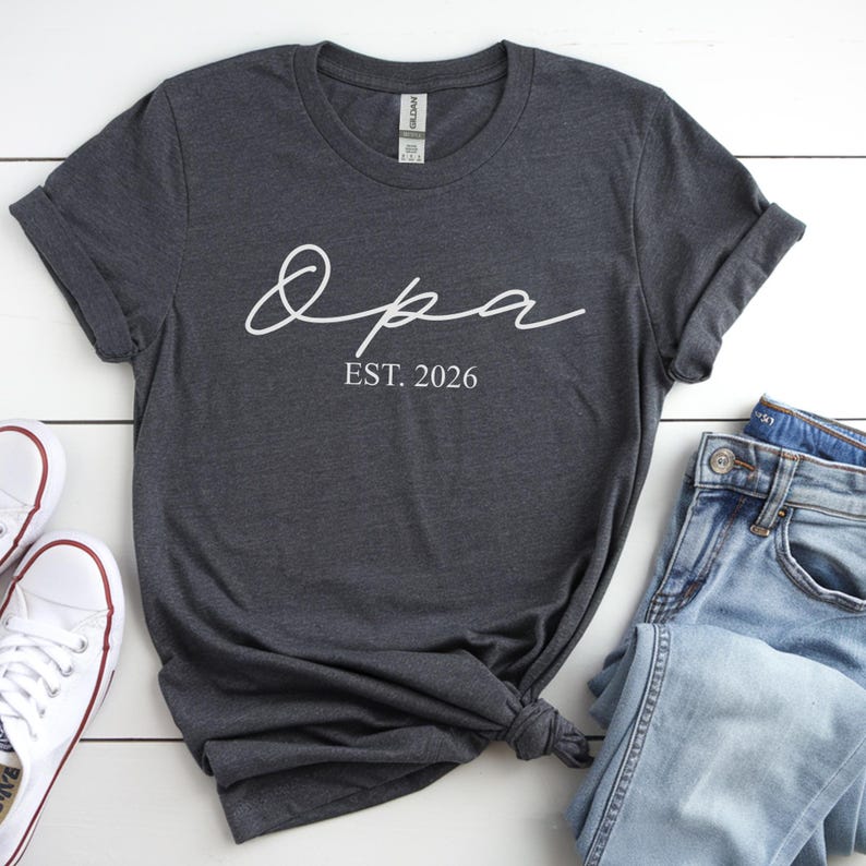 May include: Dark gray t-shirt with the word "Opa" in a white script font, and the text "EST. 2026" below it. The shirt is styled with rolled-up sleeves, a pair of jeans, and white sneakers.