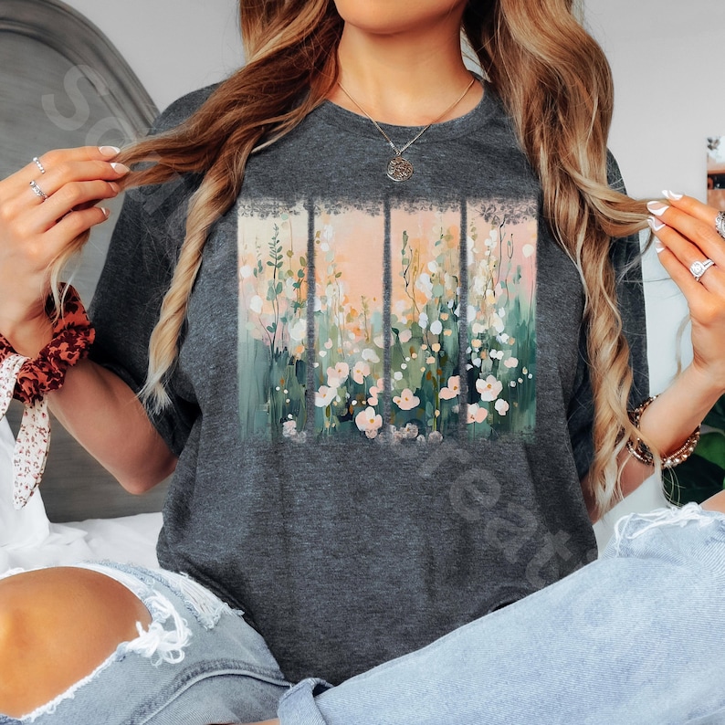 May include: Dark gray t-shirt featuring a floral design. The design is divided into four vertical panels with a watercolor-style illustration of white and pink flowers, green foliage, and a peach-colored background. The shirt is a crew neck.