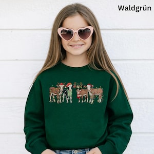 Può includere: Una felpa verde scuro con un design festivo di mucche vestite con abiti natalizi, tra cui cappelli di Babbo Natale e corna di renna. La parola "Waldgrün" è stampata in bianco. La persona indossa occhiali da sole a forma di cuore.