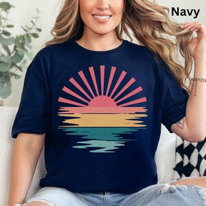 May include: Navy blue t-shirt featuring a retro sunset design. The graphic includes a pink and red sun with yellow, pink, and teal stripes below, resembling a sunset over water. The word "Navy" is printed in the upper right corner.