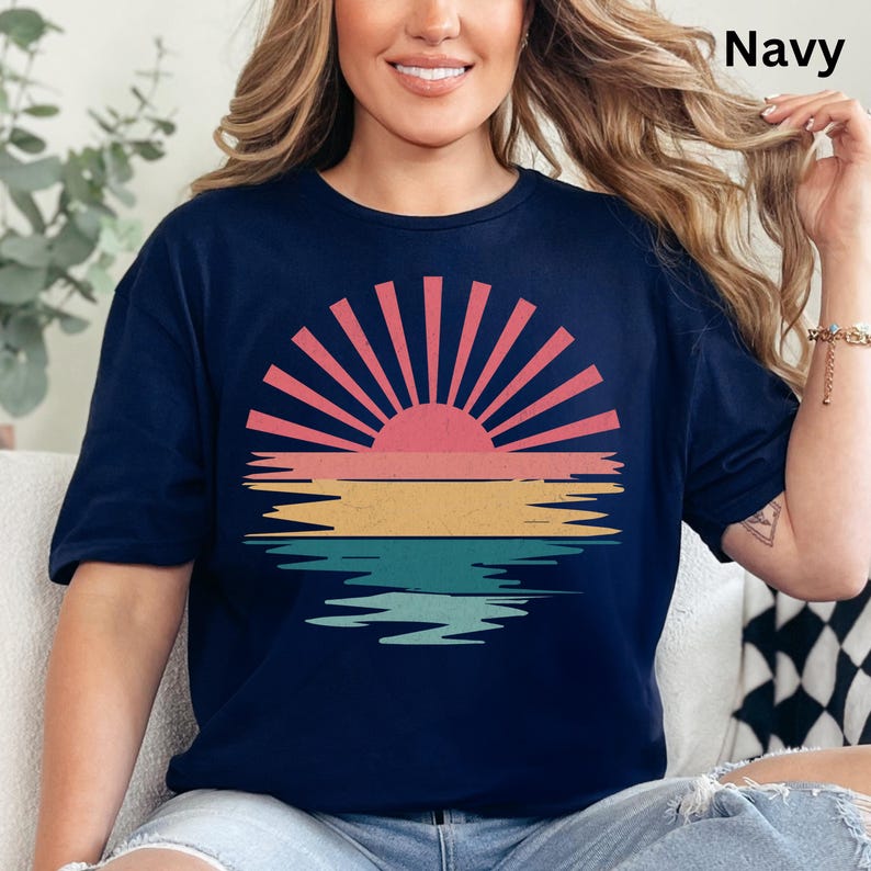 May include: Navy blue t-shirt featuring a retro sunset design. The graphic includes a pink and red sun with yellow, pink, and teal stripes below, resembling a sunset over water. The word "Navy" is printed in the upper right corner.