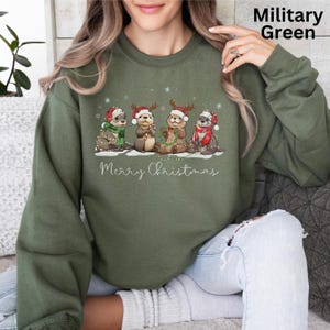Pu&ograve; includere: Felpa verde militare con un design natalizio. La grafica mostra quattro lontre con cappelli e sciarpe di Babbo Natale, con la scritta "Merry Christmas" sotto. Le lontre sono decorate con luci natalizie e tengono bastoncini di zucchero.