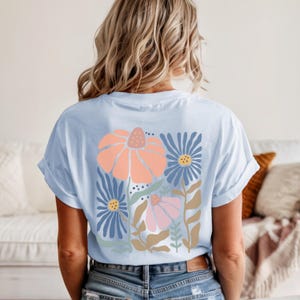 May include: Light blue t-shirt featuring a floral design on the back. The design includes a large peach-colored flower, blue flowers, and smaller pink flowers with green and brown accents. The sleeves are rolled up.
