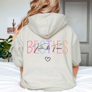May include: A cream-coloured hoodie featuring the word "BESTIES" in a rainbow of colours on the back. Below, the names "Kathi & Franzi" are written in a script font, with a small heart graphic.
