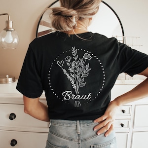 Puede incluir: Camiseta negra con un dise&ntilde;o floral blanco y la palabra "Braut" en escritura cursiva. El dise&ntilde;o incluye un coraz&oacute;n y un c&iacute;rculo punteado. La camiseta est&aacute; hecha de un material suave.