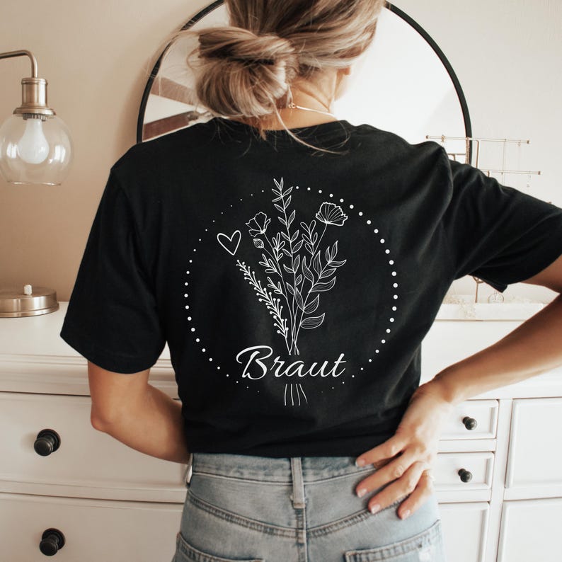 Puede incluir: Camiseta negra con un dise&ntilde;o floral blanco y la palabra "Braut" en escritura cursiva. El dise&ntilde;o incluye un coraz&oacute;n y un c&iacute;rculo punteado. La camiseta est&aacute; hecha de un material suave.
