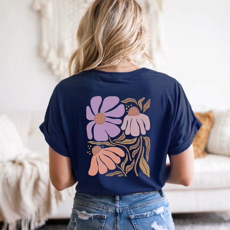 May include: A navy blue short-sleeved t-shirt featuring a large graphic print on the back. The design displays three stylized flowers in shades of light purple, dusty pink, and peach, accented with brown stems and leaves on a dark blue background.
