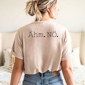 Può includere: T-shirt beige con maniche arrotolate e la scritta nera "Ähm. NÖ.". La maglietta è realizzata in materiale morbido e viene indossata con jeans blu. La maglietta è un capo casual.