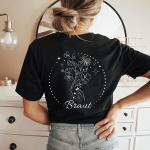 Pu&ograve; includere: T-shirt nero con un disegno floreale bianco sul retro. Il disegno presenta una mano che tiene dei fiori all'interno di un cerchio punteggiato, con la parola "Braut" sotto. La maglietta &egrave; a maniche corte e in materiale morbido.