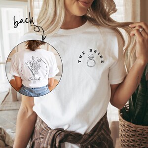 Op de afbeelding: Wit T-shirt met de tekst "THE BRIDE" en een diamantring-afbeelding. De achterkant van het shirt heeft een bloemenontwerp met de naam "Sarah". Het shirt is een casual stijl, perfect voor een bruiloftsevenement.