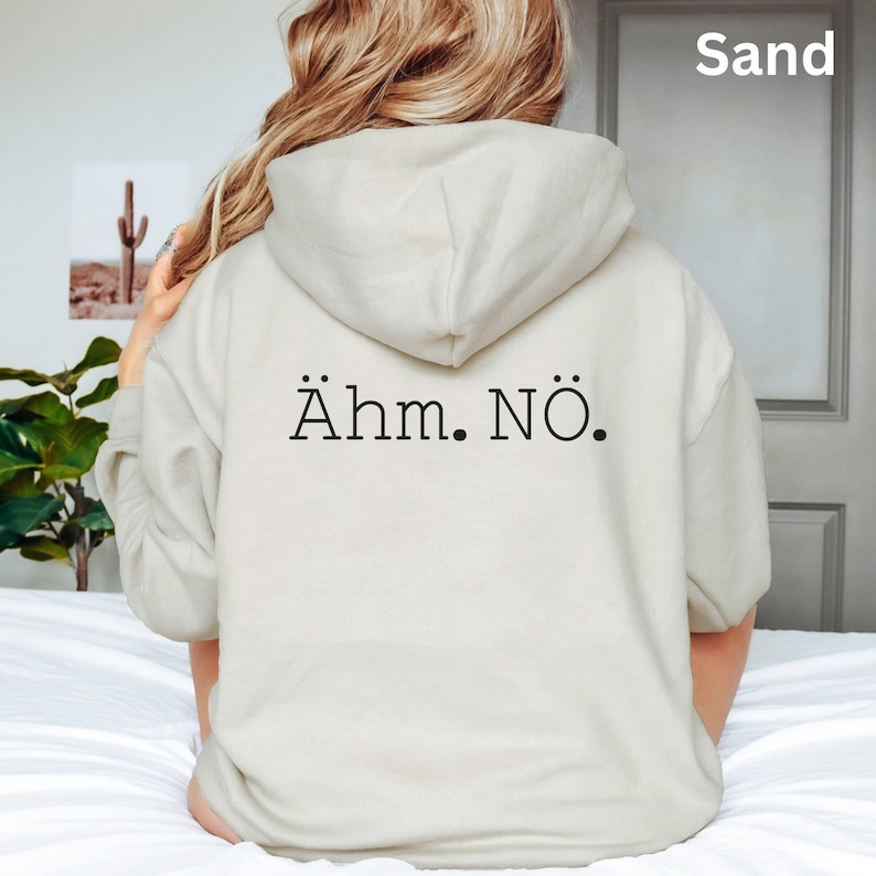 以下が含まれることがあります： 背中に黒で「&Auml;hm. N&ouml;.」とプリントされたサンドカラーのパーカー。フードとゆったりとしたフィット感が特徴です。「Sand」の文字が右上に白でプリントされています。