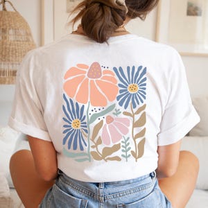 May include: White t-shirt featuring a colorful floral design on the back. The design includes large peach and blue flowers, with smaller pink flowers and green leaves. The shirt is made of a soft material and is suitable for casual wear.