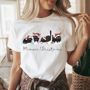 May include: White t-shirt featuring a festive design with four black cats wearing Santa hats and reindeer antlers. The cats are surrounded by snowflakes and Christmas lights, with the text "Meowee Christmas" below.