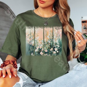 May include: An olive green t-shirt featuring a floral design. The design is a rectangular panel divided into four vertical sections, with a soft pink and white floral pattern. The shirt has a crew neck and short sleeves.