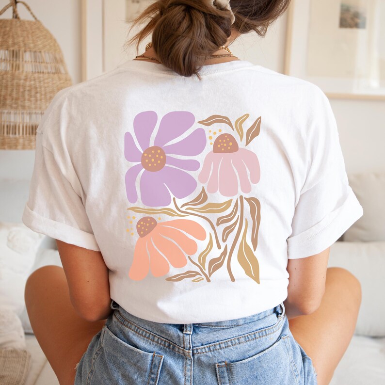 May include: A white short-sleeved t-shirt featuring a large graphic on the back. The design consists of three stylized flowers in shades of lavender, light pink, and peach, accompanied by tan-colored leaves and stems on a white background.