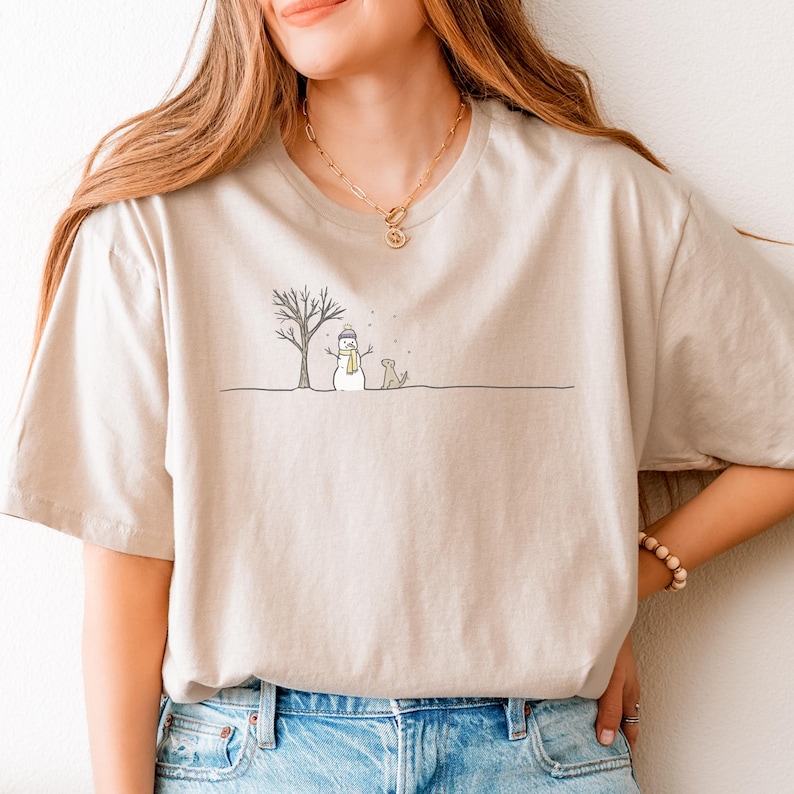 May include: A beige short-sleeve t-shirt featuring a minimalist line art graphic of a snowman wearing a blue hat and yellow scarf, a bare tree, and a dog standing in the snow. The design is centered on the chest of the crew neck garment.