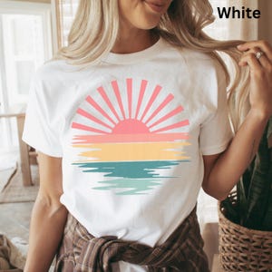 May include: White t-shirt featuring a colorful sunset design. The graphic includes a pink sun with radiating lines, above horizontal bands of yellow, teal, and orange, with a reflection below. The word "White" is printed on the top right.