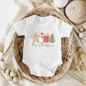 May include: White baby bodysuit with a festive gingerbread man, snowman, gift, and Christmas tree design. The words "Merry Christmas" are written in a script font below the illustrations. The bodysuit is displayed in a woven basket.
