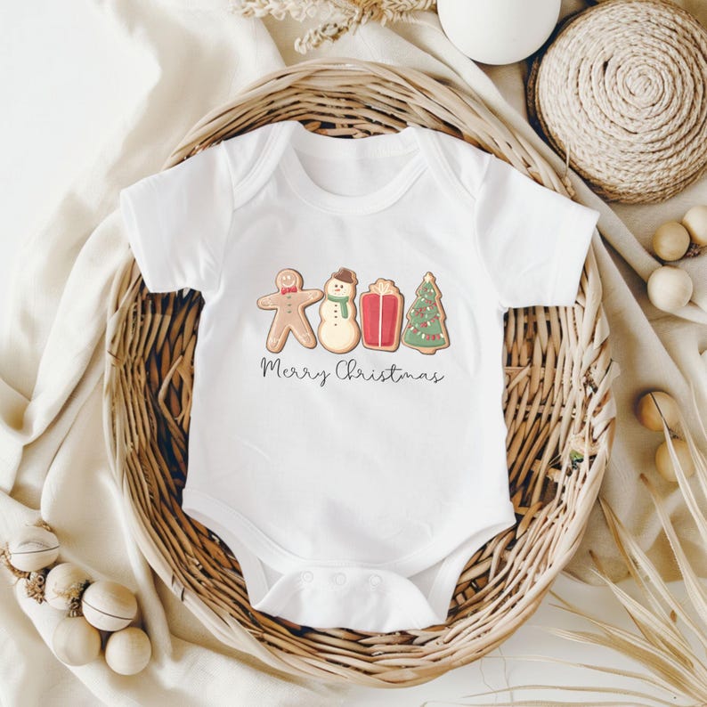 May include: White baby bodysuit with a festive gingerbread man, snowman, gift, and Christmas tree design. The words "Merry Christmas" are written in a script font below the illustrations. The bodysuit is displayed in a woven basket.