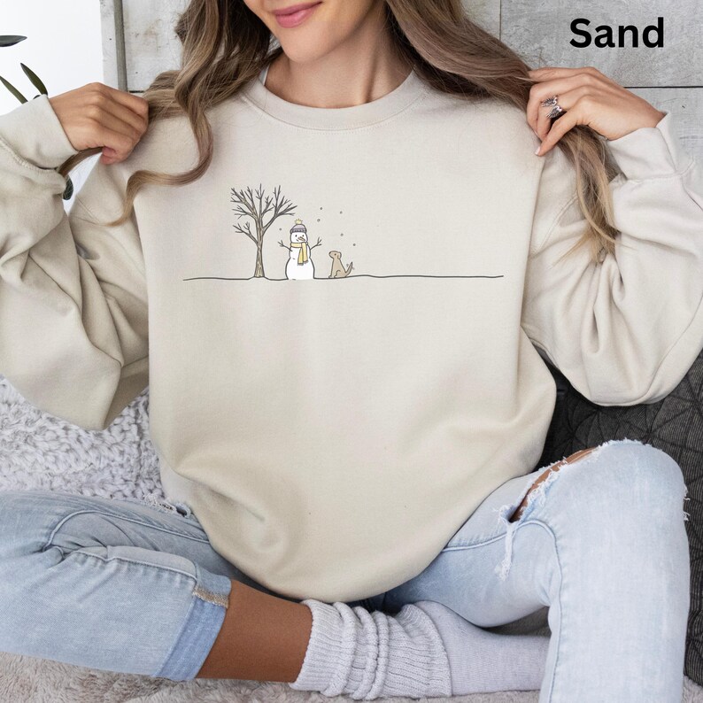May include: A sand-colored crewneck sweatshirt featuring a minimalist line art graphic of a snowman wearing a yellow scarf, a bare tree, and a dog standing on a horizontal line, with the word Sand printed in the upper right corner.