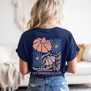 May include: A navy blue t-shirt featuring a floral design in peach, blue, and pink. The graphic includes stylized flowers and the text "LET KINDNESS BLOOM" in white. The shirt is a casual style.