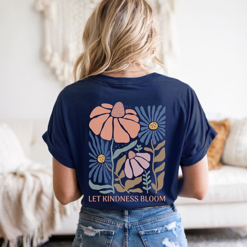 May include: A navy blue t-shirt featuring a floral design in peach, blue, and pink. The graphic includes stylized flowers and the text "LET KINDNESS BLOOM" in white. The shirt is a casual style.