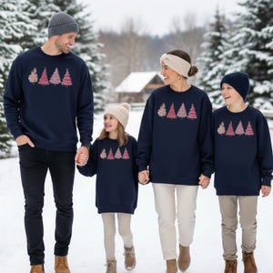 K&ouml;nnte beinhalten: Eine Familie tr&auml;gt passende marineblaue Sweatshirts mit einem festlichen Weihnachtsbaumdesign. Die Sweatshirts zeigen eine Reihe von vier rosa und goldenen Weihnachtsb&auml;umen. Die Familie geht in einer verschneiten Winterlandschaft spazieren.