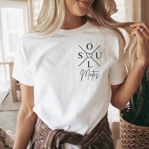 May include: White t-shirt featuring the words "SOUL" and "MATES" in black. "SOUL" is in a cross design with a heart replacing the "O". "MATES" is written in cursive. A casual, everyday wear item.