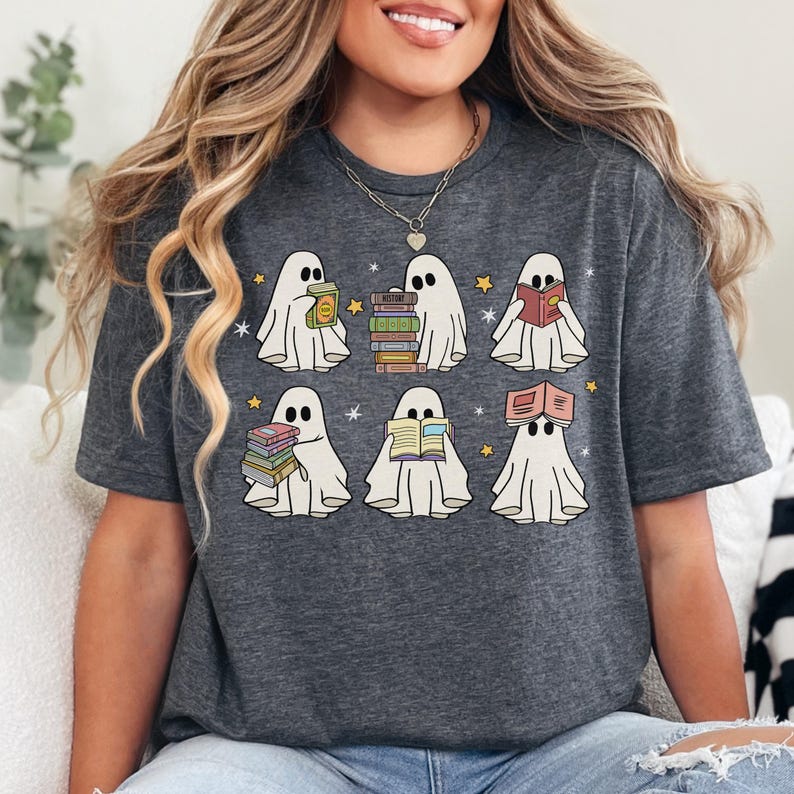 May include: Dark gray t-shirt featuring a whimsical design of six cartoon ghosts, each holding or reading books. The ghosts are white with black eyes and are surrounded by yellow stars. A fun, book-themed graphic tee.