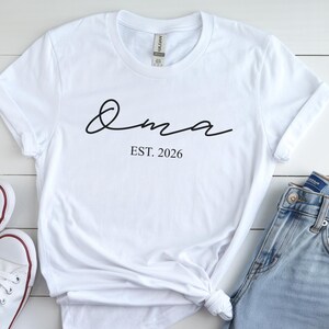 May include: White t-shirt with the word "Oma" in a cursive font and the text "EST. 2026" in a simple font. The shirt is styled with rolled-up sleeves and a knot at the bottom.