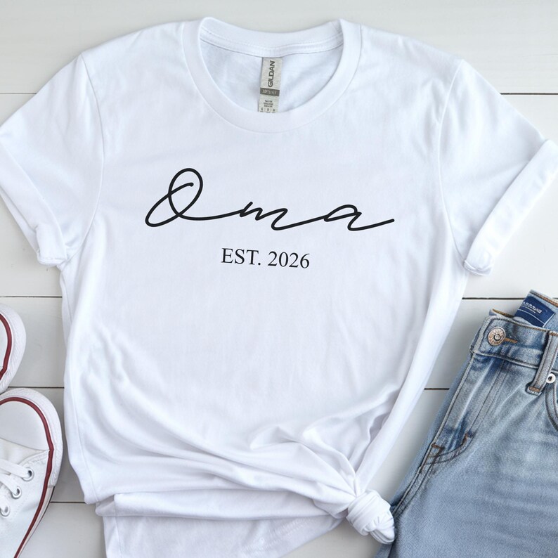 May include: White t-shirt with the word "Oma" in a cursive font and the text "EST. 2026" in a simple font. The shirt is styled with rolled-up sleeves and a knot at the bottom.