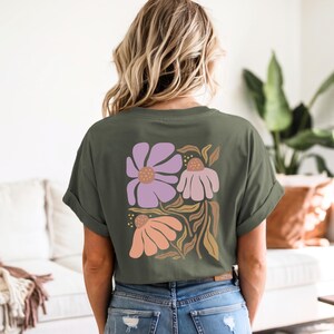 May include: An olive green short-sleeve t-shirt featuring a large graphic on the back. The design displays three stylized flowers in shades of lavender, light pink, and peach, accompanied by tan-colored leaves and stems on a dark green background.