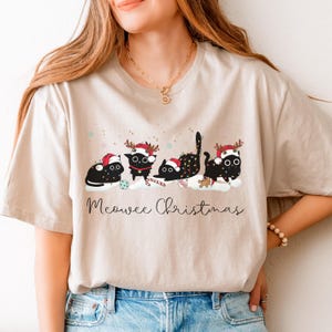 May include: Beige t-shirt featuring a festive design with four black cats wearing Santa hats and reindeer antlers. The cats are decorated with Christmas lights and candy canes, with the text "Meowee Christmas" below.