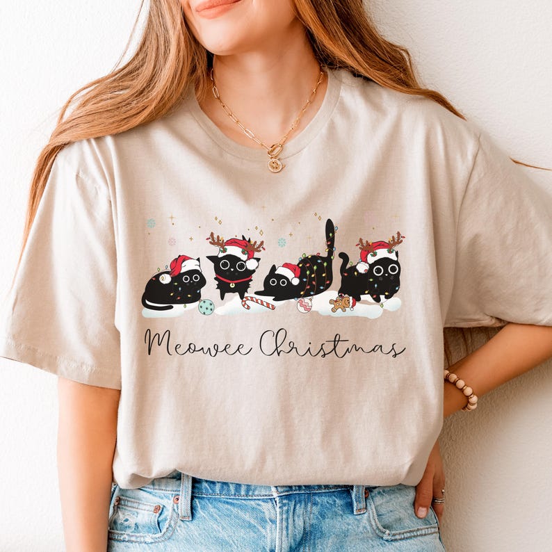 May include: Beige t-shirt featuring a festive design with four black cats wearing Santa hats and reindeer antlers. The cats are decorated with Christmas lights and candy canes, with the text "Meowee Christmas" below.