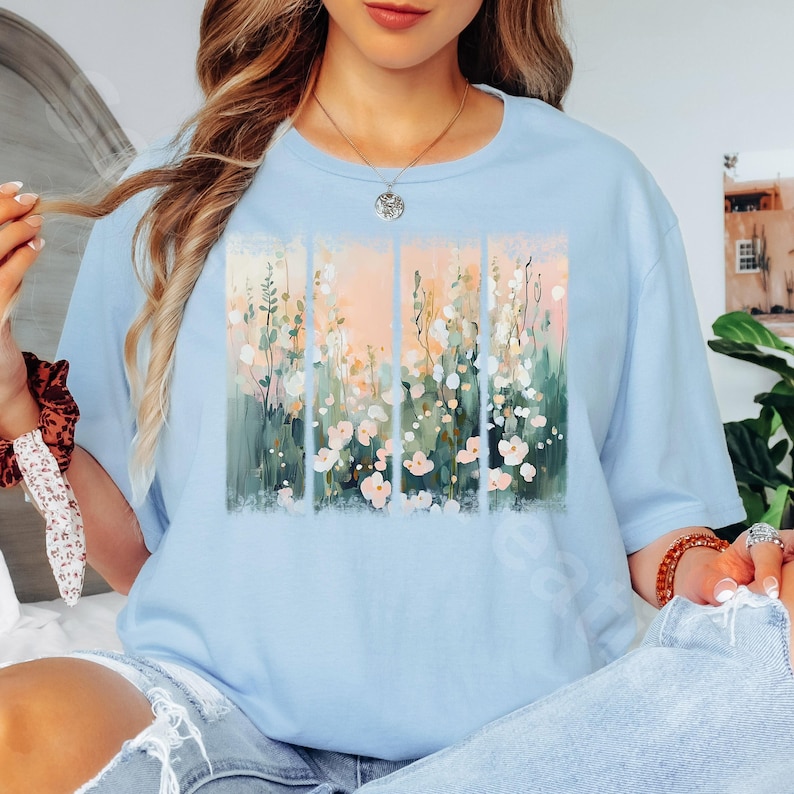 May include: A light blue t-shirt featuring a floral design. The artwork showcases vertical panels with a pastel color palette of pink, green, and white flowers. The shirt has a crew neck and short sleeves, perfect for casual wear.