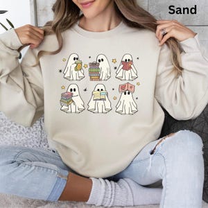 May include: Sand-colored sweatshirt featuring a graphic of six cartoon ghosts reading books. Each ghost is depicted with a white sheet and black eyes, surrounded by stars. The word "Sand" is printed in the upper right corner.