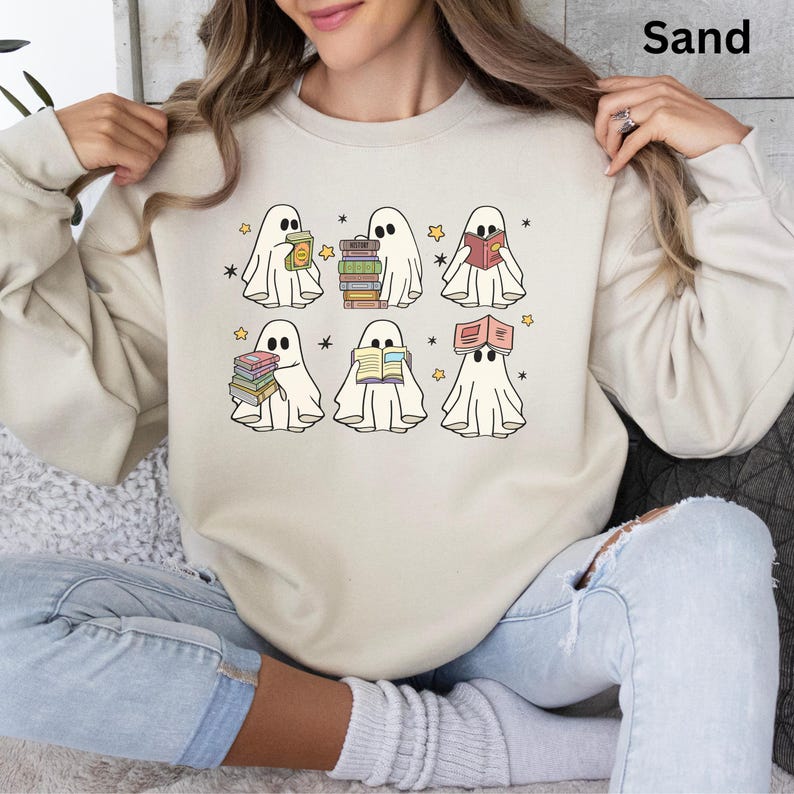 May include: Sand-colored sweatshirt featuring a graphic of six cartoon ghosts reading books. Each ghost is depicted with a white sheet and black eyes, surrounded by stars. The word "Sand" is printed in the upper right corner.