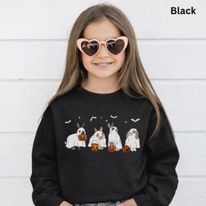 Op de afbeelding: Zwart sweatshirt met een Halloween-ontwerp. Het ontwerp toont vier konijnen in spookkostuums, elk met een pompoenemmer. Het woord "Black" staat rechtsboven afgedrukt. Het meisje op de foto draagt hartvormige zonnebrillen.