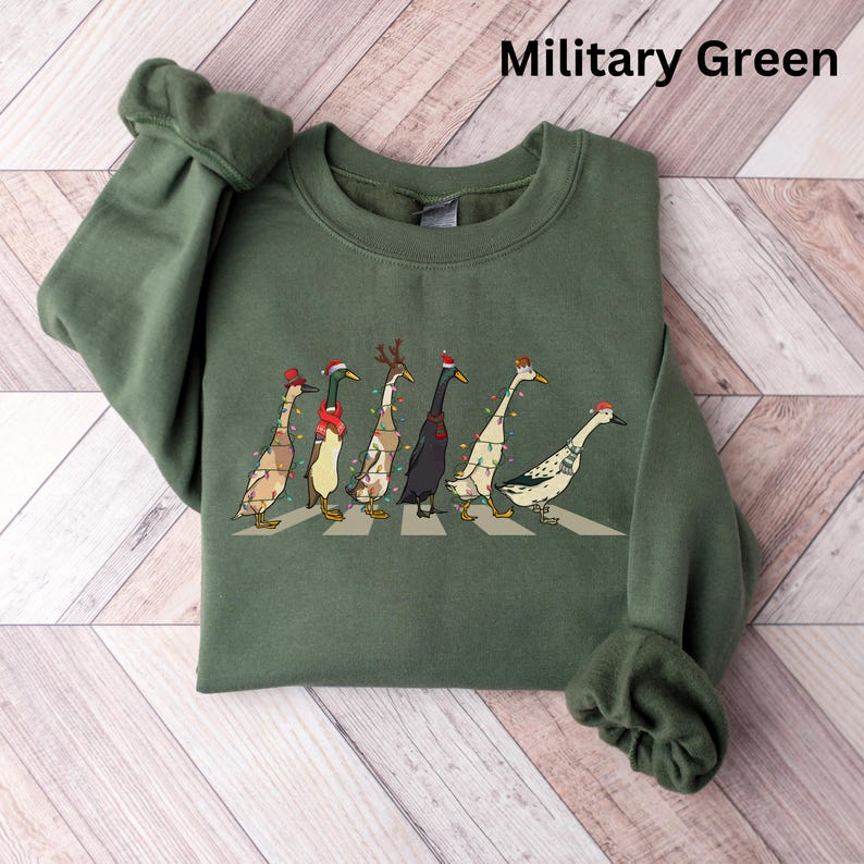 Peut inclure: Un sweat-shirt vert avec un motif de No&euml;l repr&eacute;sentant une ligne de canards portant des chapeaux de P&egrave;re No&euml;l et des lumi&egrave;res de No&euml;l. Le texte "Military Green" est affich&eacute; en haut de l'image.
