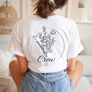 Puede incluir: Camiseta blanca con un dise&ntilde;o floral negro en la espalda, que incluye un coraz&oacute;n y la palabra "Crew" en cursiva. El dise&ntilde;o est&aacute; encerrado en un c&iacute;rculo punteado. La persona lleva pantalones cortos vaqueros azules.