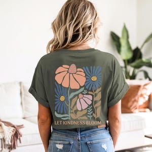 May include: Olive green t-shirt with a floral design on the back. The design features orange, blue, and pink flowers with the text "LET KINDNESS BLOOM" below the flowers. The shirt has rolled-up sleeves.