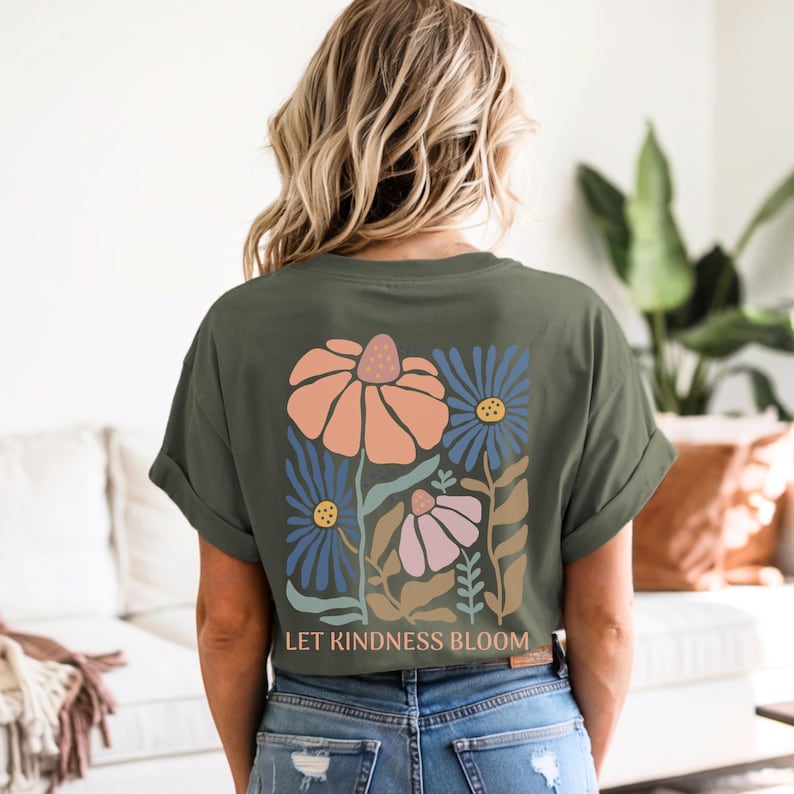 May include: Olive green t-shirt with a floral design on the back. The design features orange, blue, and pink flowers with the text "LET KINDNESS BLOOM" below the flowers. The shirt has rolled-up sleeves.