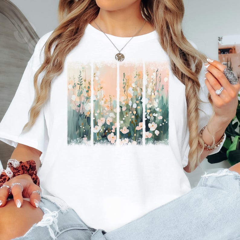 May include: White t-shirt featuring a floral design. The artwork showcases a pastel-colored meadow with pink and white flowers, green foliage, and a soft peach-colored sky. The design is divided into vertical panels.