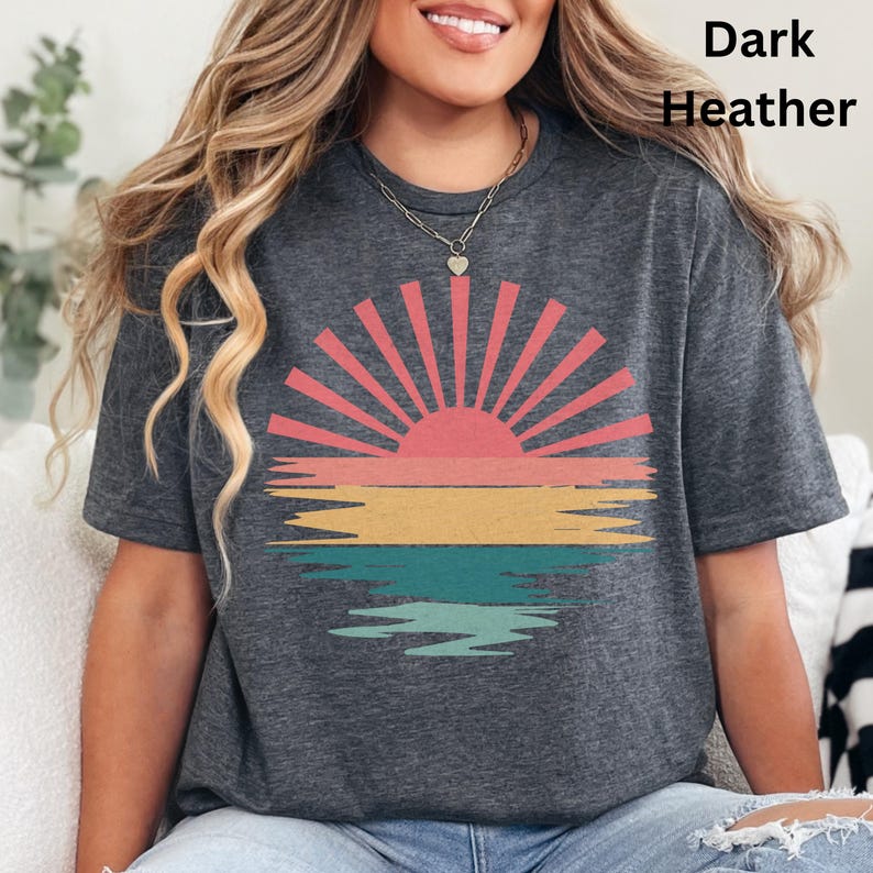 May include: Dark heather gray t-shirt featuring a retro sunset design. The graphic includes a pink and orange sun with rays, above horizontal stripes of pink, yellow, and teal. The text "Dark Heather" is printed in the upper right corner.