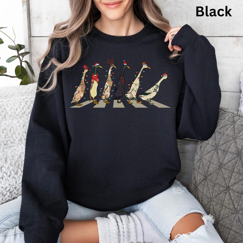 Peut inclure: Sweat-shirt noir avec un motif de canards portant des lumi&egrave;res de No&euml;l et des chapeaux marchant sur un passage pour pi&eacute;tons. Le texte "Black" est visible dans le coin sup&eacute;rieur droit de l'image.