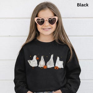 Peut inclure: Sweat-shirt noir avec une rangée de fantômes de dessins animés tenant des citrouilles. Les fantômes sont blancs avec des ombres grises, et les citrouilles sont oranges. Le mot "Black" est imprimé en haut à droite.