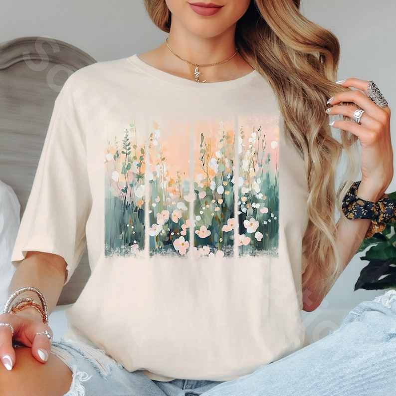 May include: Cream-colored t-shirt featuring a floral design. The artwork showcases a vertical arrangement of flowers in shades of pink, white, green, and peach. The shirt has a classic crew neck and short sleeves, suitable for casual wear.